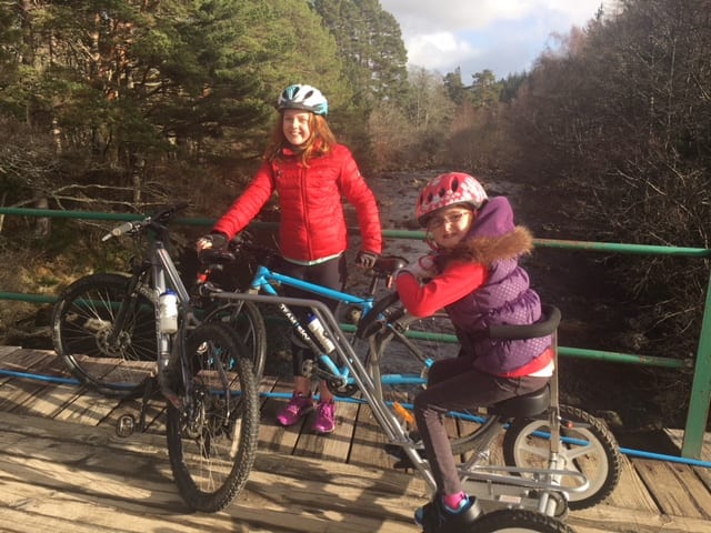Isla sitting on a bike, on a bridge with the river behind